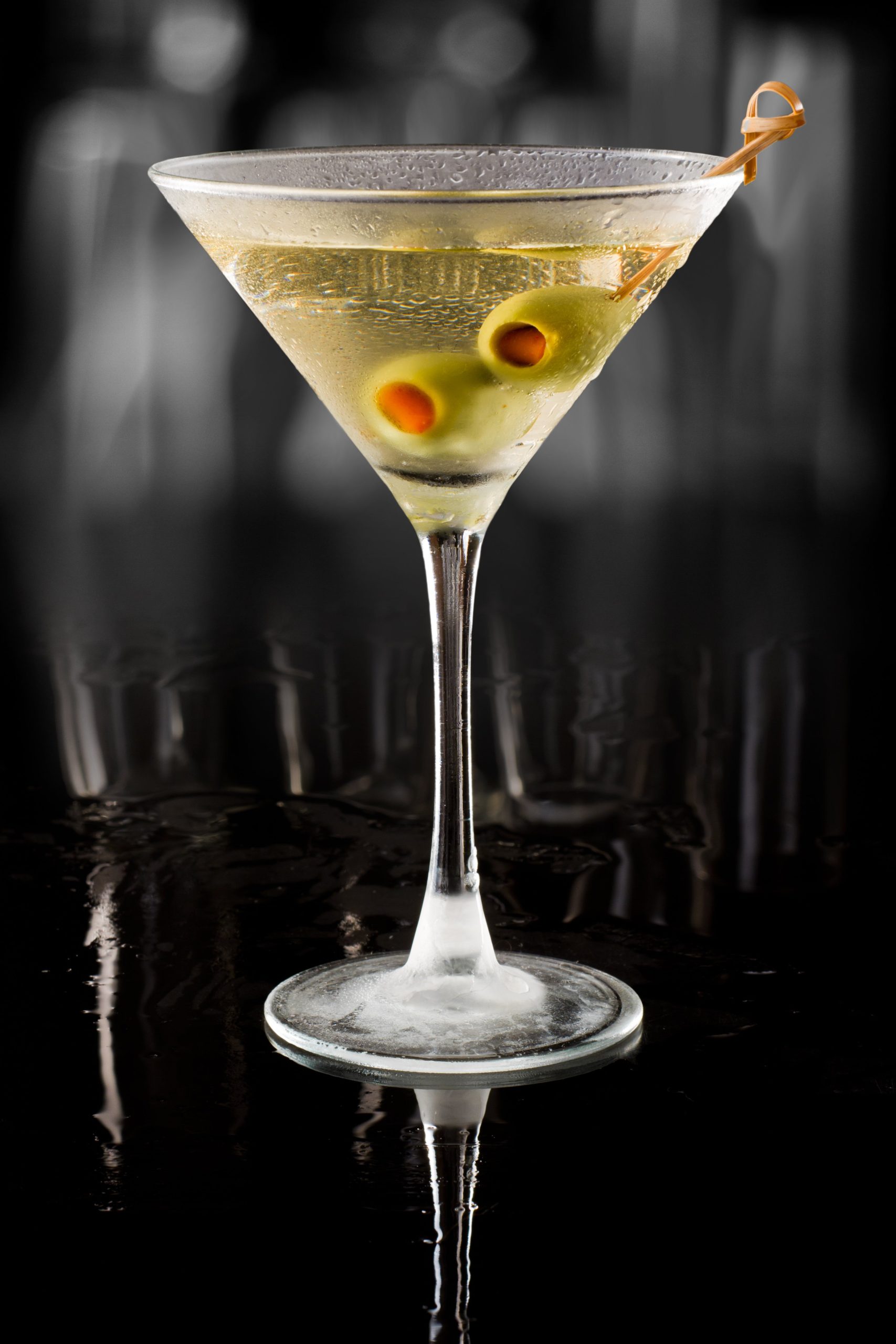 Martini glass with a clear cocktail garnished with green olives on a toothpick, set against a dark background